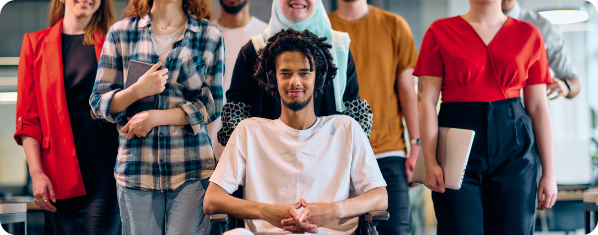 A diverse group of young people standing together, with a man in a wheelchair at the center, smiling confidently. They are dressed in casual attire and appear to be in a modern office environment.