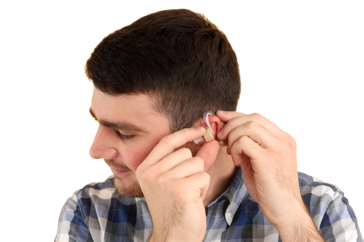 A man wearing a plaid shirt is putting a hearing aid into his ear, smiling slightly.