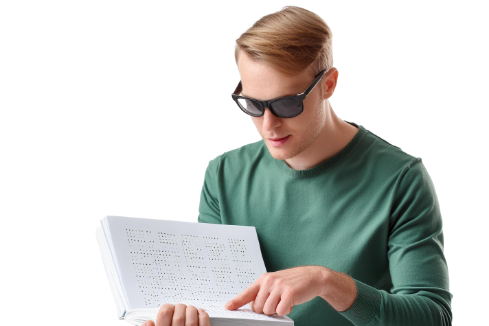 A man wearing sunglasses and a green sweater reads a Braille book, using his finger to follow the text.