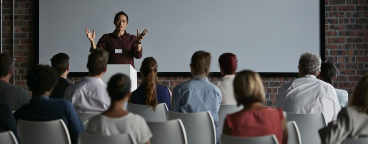 A person is presenting at a podium in front of an audience in a conference room with a large screen.