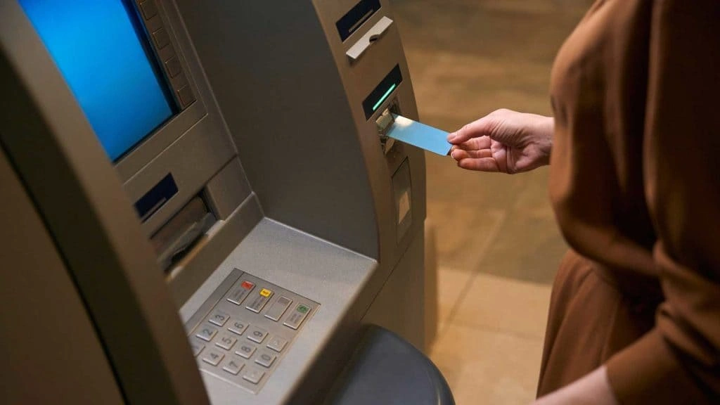شخص في مكان داخلي يستخدم بطاقة ائتمان لإجراء عملية دفع في ماكينة صرف آلي، محاطًا بلوازم مكتبية وحائط في الخلفية.