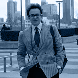A man in a checkered suit and glasses stands confidently on a city sidewalk, with skyscrapers behind him.