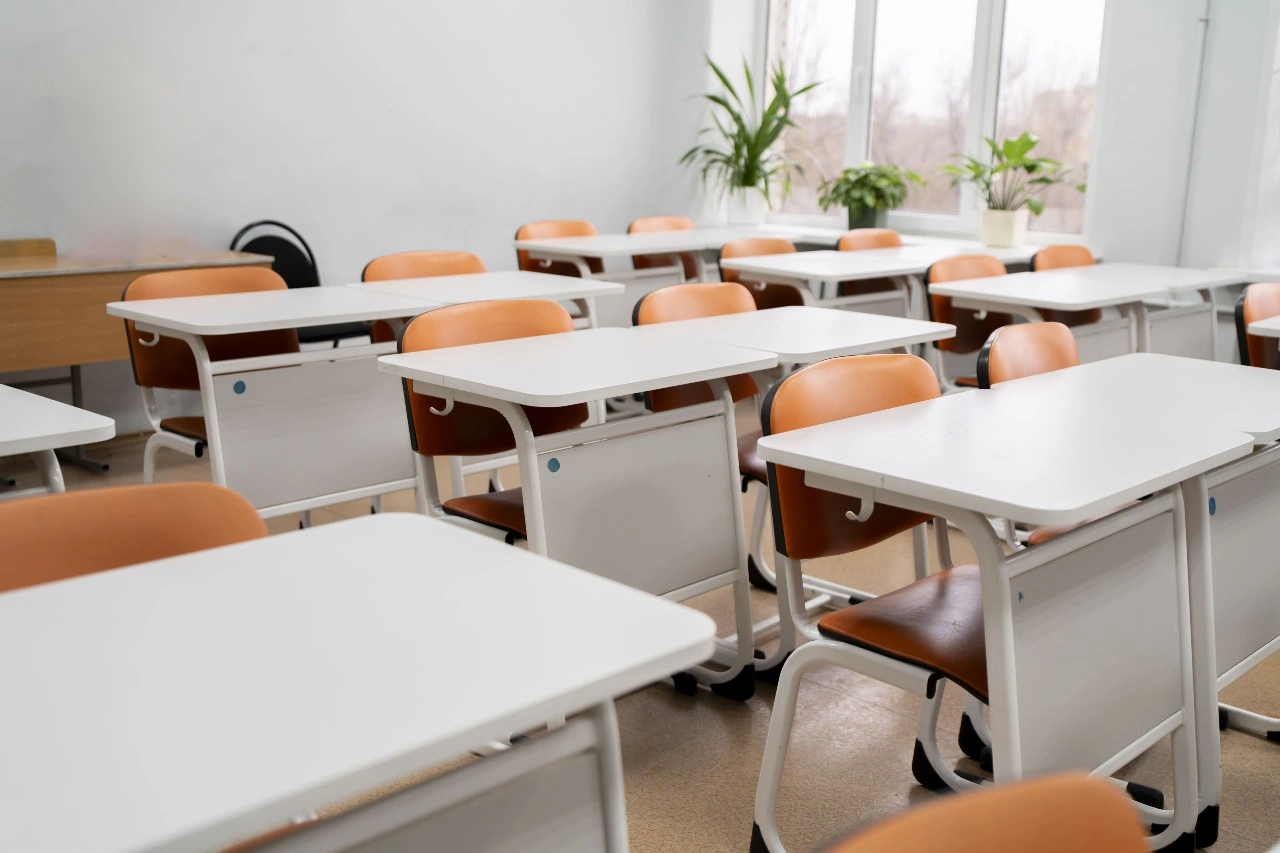 فصل دراسي يضم العديد من المكاتب والكراسي الموزعة في جميع أنحاء المكان، مع وجود لوحة بيضاء على أحد الجدران وأرضية ناعمة.