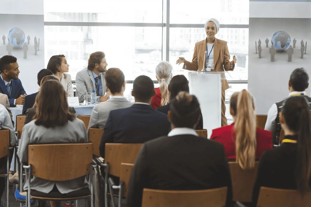 امرأة تقف أمام مجموعة من الأشخاص في قاعة دراسية، مع وجود السبورة البيضاء والأثاث المرئي.