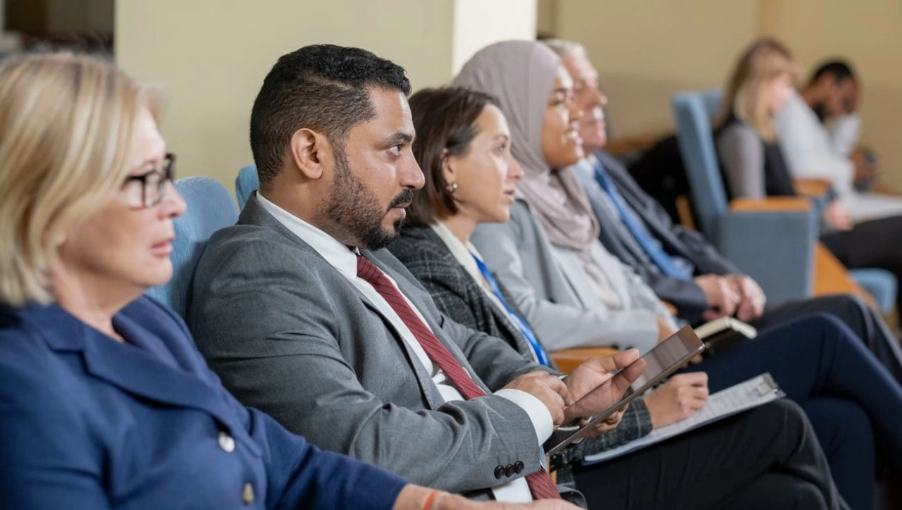 مجموعة من الأفراد المتنوعين، رجالاً ونساءً، يجلسون على كراسي في قاعة اجتماعات. يرتدون ملابس رسمية، بما فيها البدلات. يسود جو من التعاون والنقاش بين أفراد المجموعة أثناء تفاعلهم مع بعضهم البعض.