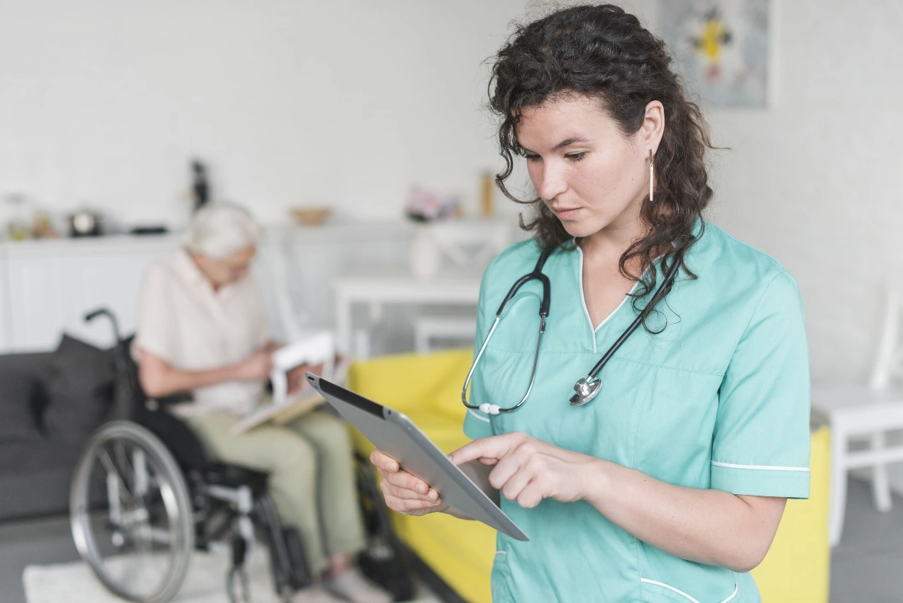 A woman wearing scrubs is using a tablet while sitting in a hospital environment. There is medical equipment nearby and a wheelchair in the background.