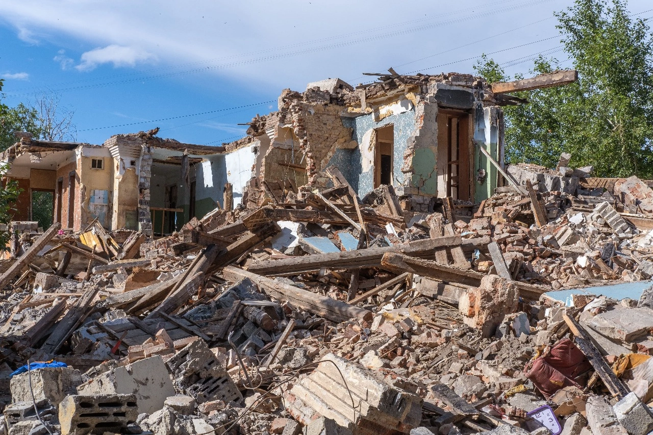 A building surrounded by debris and rubble, with scattered litter and destruction in the outdoor environment under a cloudy sky. Nearby, there are trees amidst the chaos of a natural disaster.