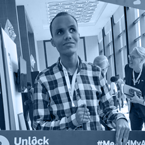 A person in a checkered shirt and lanyard holds a frame at an event, surrounded by attendees in a well-lit room.