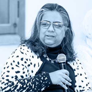 A woman speaks into a microphone, dressed in a black top and a black-and-white spotted cardigan.