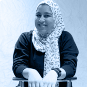 A smiling person in a patterned hijab and black top leans on a chair, radiating a friendly and relaxed vibe.