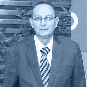 A man wearing a formal suit and tie stands against a backdrop of greenery and wooden panels.