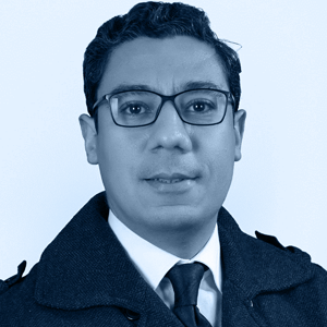 A man in a formal coat and tie stands against a soft blue background, radiating professionalism and sophistication.