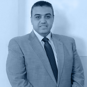 A formally dressed man in a gray suit and tie stands against a light wall, exuding a professional demeanor.