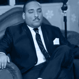 A man in a suit sits relaxed on a leather couch, eyes closed, with a patterned wallpaper and lamp in the background.