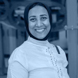 A smiling woman wearing a black hijab and a white shirt is standing indoors with a mechanical background, giving off a warm and welcoming atmosphere.