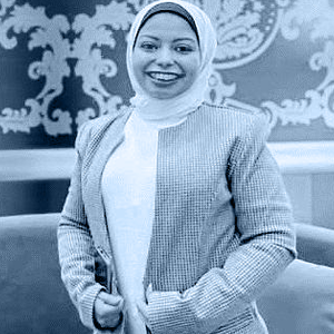 A woman wearing a headscarf smiles indoors, with a wall in the background.