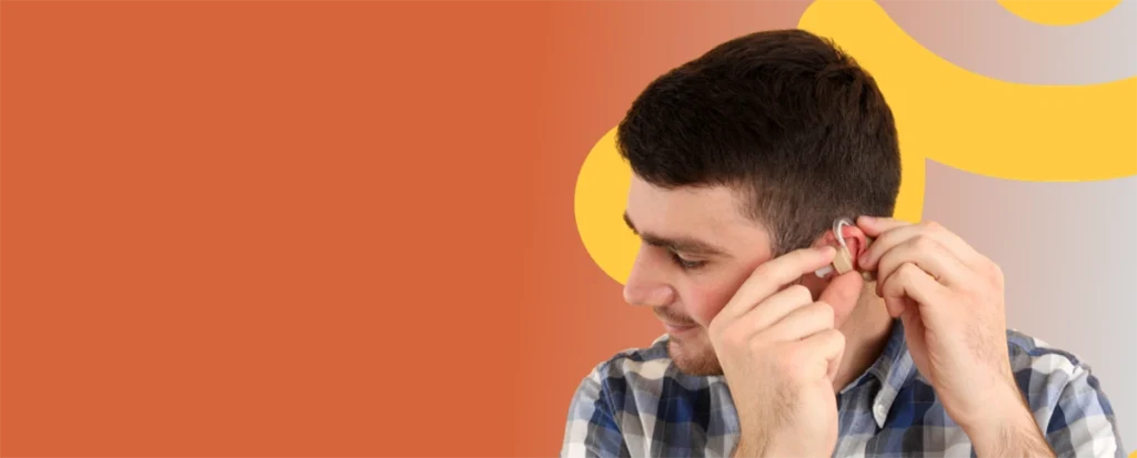 A man in a plaid shirt adjusts his hearing aid, smiling, against an orange and yellow background.